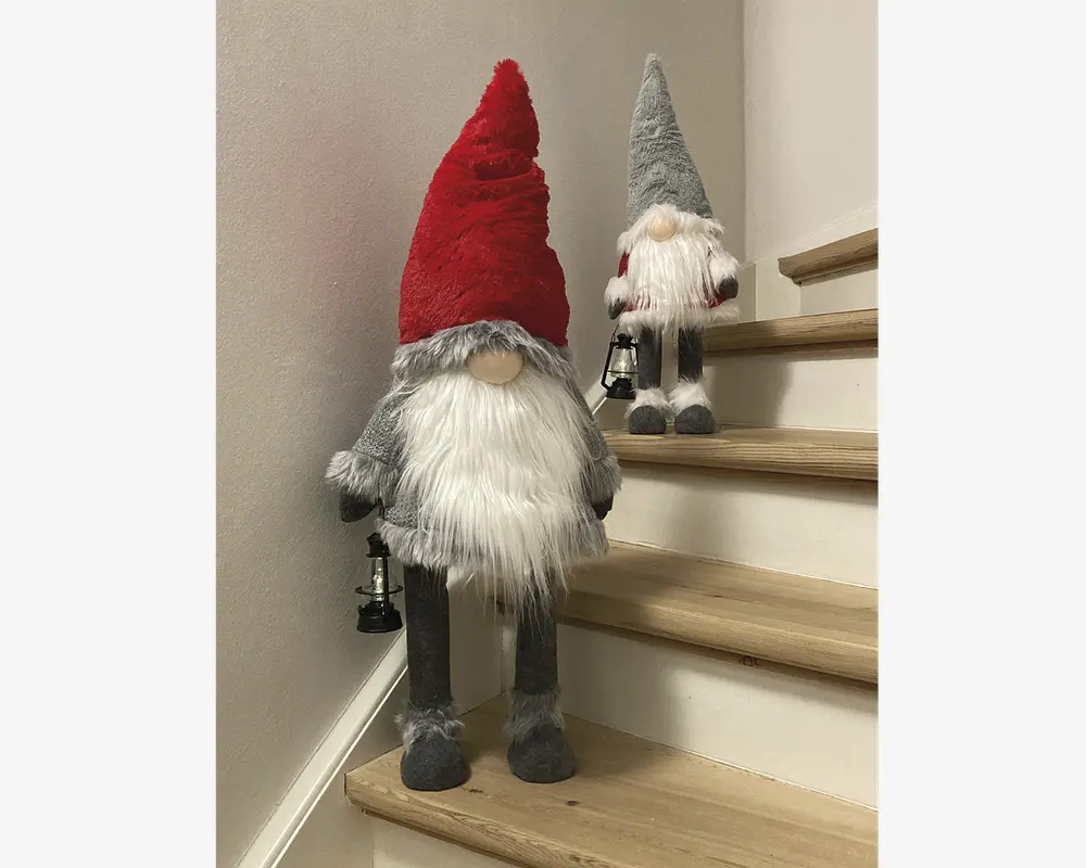 To plyssede nissefigurer med lange hvide skæg og runde næser, hver med en lille sort lanterne i hånden. Den største nisse har en høj, spids rød hue, gråt tøj og står på det nederste trappetrin, mens den mindre nisse har en høj, spids grå hue, hvidt skæg og står på et højere trin. Begge har bløde, lodne teksturer og er designet til at stå oprejst på flade overflader.