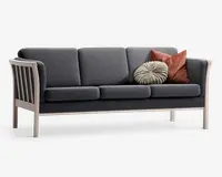 En moderne tresæders sofa med mørkegråt stofbetræk og stel i lyst træ. Sofaen har tre sædehynder og tre ryghynder, alle med rene, lige linjer. Armlæn og ben er lavet af lyst træ med lameller i siderne for et moderne udtryk. Der medfølger to pyntepuder: én rund, plisseret beige pude og én firkantet, rustfarvet pude.