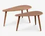 Sæt med to borde i træ med et design inspireret af midten af det 20. århundrede. Hvert bord har en glat, dråbeformet bordplade og tre skråtstillede, koniske ben. Træet har en naturlig finish, der fremhæver årestrukturen. Bordene er i forskellige størrelser, så de kan placeres sammen eller delvist ind under hinanden for et lagdelt udtryk.