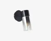 Et moderne vægmonteret armatur med en cylindrisk klar glasskærm og sort metalbase. Armaturet har en synlig pære indeni, og designet er slankt og minimalistisk, velegnet til moderne indretning.