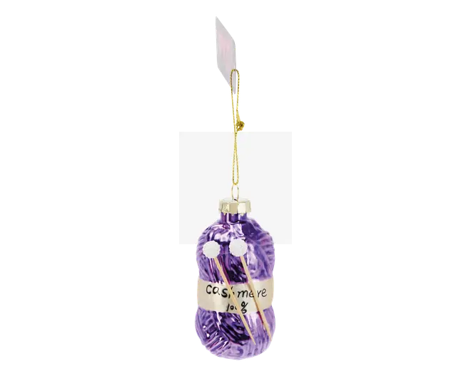 En dekorativ pynt formet som et garnnøgle i lilla med en cremefarvet etiket, hvor der står 'cashmere 100%'. Ornamentet har to små hvide pomponer og to miniaturestrikkepinde stukket ind i garnet. Det hænger i en guldfarvet glitrende snor med et hvidt mærke øverst.
