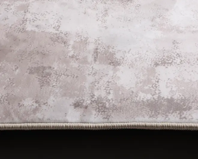 Nærbillede af en blød, plysset overflade med et diskret abstrakt mønster i lysegrå og beige nuancer. Kanten er afsluttet med en pænt syet, lys kant, hvilket giver et rent og holdbart udseende.