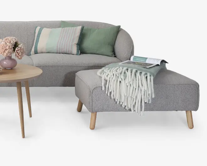 Et moderne, lysegråt polstret siddemøbel med afrundede kanter og ben i lyst træ. Sættet inkluderer en matchende skammel, der er pyntet med et blødt, mintgrønt plaid med frynser og et åbent magasin ovenpå. Sofaen er dekoreret med tre pyntepuder i afdæmpede grønne og stribede pastelfarver. Et rundt sidebord i træ har en lille vase med lyserøde blomster, hvilket giver et hyggeligt og indbydende udtryk.