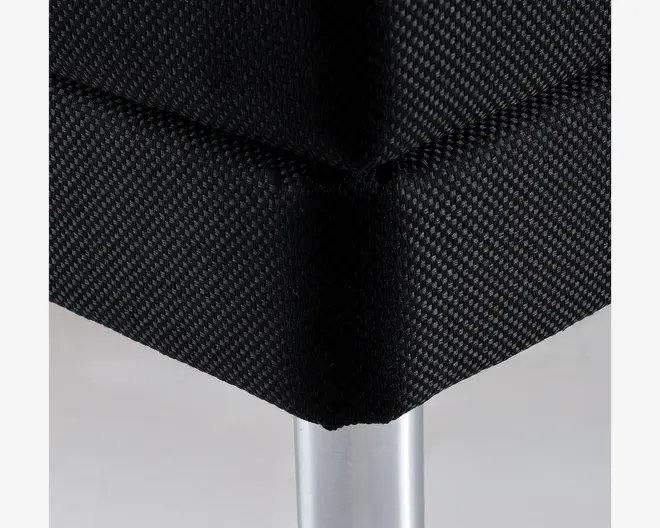 Nærbillede af et møbelhjørne med tæt vævet sort stofbetræk og et cylindrisk sølvfarvet metalben. Syninger og tekstur i stoffet er tydeligt synlige, hvilket indikerer et holdbart og moderne design.