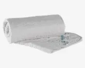 Hvid, quiltet madrasbeskytter med diskret bølget syning. Stoffet ser blødt og polstret ud for ekstra komfort. Kanterne er pænt afsluttet med en syet kant, og et produktmærke med vaskeanvisninger og certificeringer er synligt i det ene hjørne. Beskytteren er delvist rullet sammen, hvilket viser dens fleksibilitet og tykkelse.