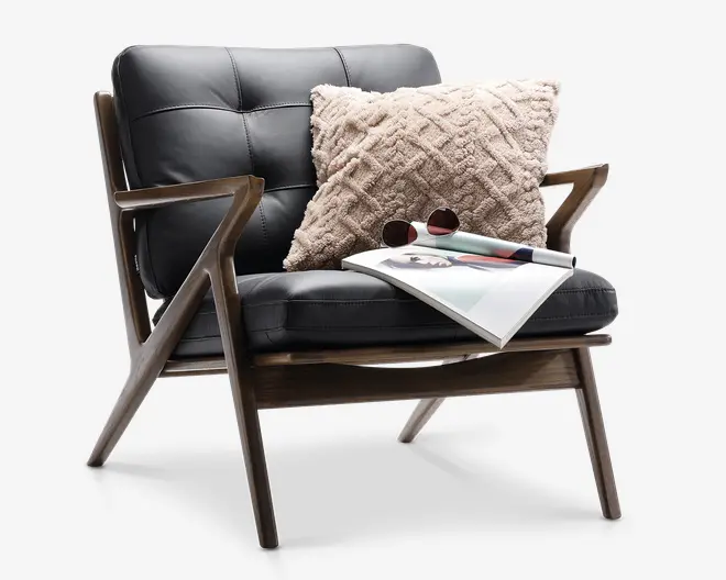 Stol med moderne, vinklet træramme og sorte, polstrede læderhynder. Indeholder en stor, beige pyntepude med diamantmønster og tekstur, samt et magasin med solbriller liggende på sædet. Stolen har brede armlæn og en lav, tilbagelænet profil.