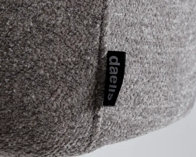 Nærbillede af en tekstureret, lysegrå stofoverflade med en synlig syning og et lille sort stofmærke med mærkenavnet 'daells' i hvide bogstaver. Stoffet ser blødt og behageligt ud, hvilket indikerer en hyggelig og komfortabel fornemmelse.