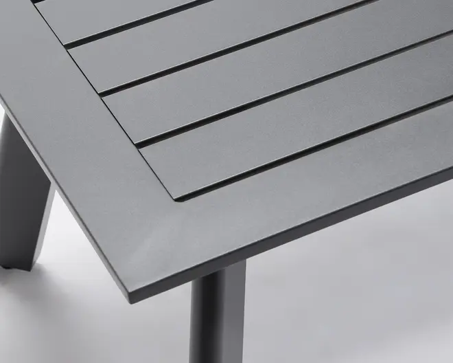 Nærbillede af et bordhjørne med mat, mørkegrå overflade. Bordpladen har et lameldesign med jævnt fordelte, vandrette paneler, og rammen er lavet af solidt metal. Benene er firkantede og robuste, hvilket giver stabil støtte.