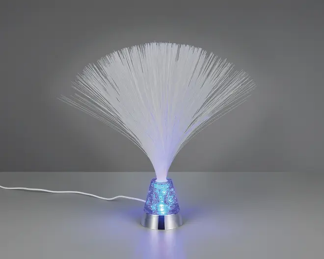 En dekorativ lampe med en sølvfarvet bund og en kegleformet, gennemsigtig blå sektion fyldt med glimmer. Tynde, hvide fiberoptiske tråde spreder sig ud fra toppen og danner en bred, fjerlignende effekt. Lampen udsender et blødt blåt skær, der oplyser fibrene og glimtet i bunden. En hvid ledning går ud fra bagsiden.