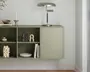 Vægmonteret skab i en afdæmpet salviegrøn farve med et minimalistisk design. Skabet har åbne hylder i venstre side, opdelt i fire rum, samt en enkelt låge til højre med en lille rund knop. Hylderne er dekoreret med genstande som en sort-hvidstribet vase, et skulpturelt keramikobjekt, en stak bøger, en hvid skål og en korallignende dekoration. Ovenpå skabet står en moderne bordlampe i metal, en høj lysestage og en lille dekorativ skål. Udtrykket er rent og moderne, velegnet til nutidige indretninger.
