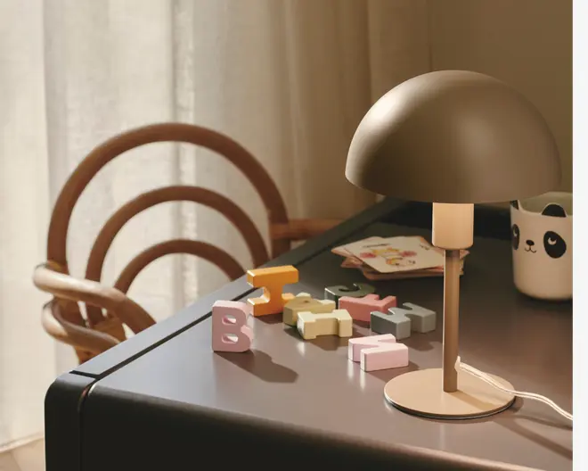 En moderne bordlampe med en mat beige, kuppelformet skærm og en slank, cylindrisk fod står på et mørkt skrivebord. Lampen har en hvid ledning. På bordet ligger pastel-farvede træbogstaver, et krus med pandaansigt og en lille illustreret notesbog. I baggrunden ses en træstol med buede ryglæn og lyse gardiner, der slipper blødt dagslys ind.