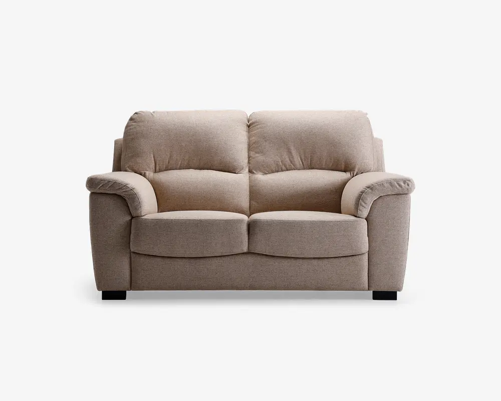 En to-personers sofa med blødt, lysebeige stofbetræk. Sofaen har fyldige, polstrede ryghynder og brede, polstrede armlæn for ekstra komfort. Sædehynderne er tykke og let buede foran, hvilket giver ergonomisk støtte. Sofaen står på korte, solide sorte ben.