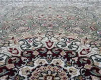 Nærbillede af et traditionelt mønstret tæppe med indviklede blomster- og geometriske motiver. Designet indeholder nuancer af grøn, creme, bordeaux og beige med detaljerede konturer i mørkegrå. Teksturen ser blød og fyldig ud, og mønsteret er symmetrisk, hvilket giver tæppet et klassisk og elegant udtryk.
