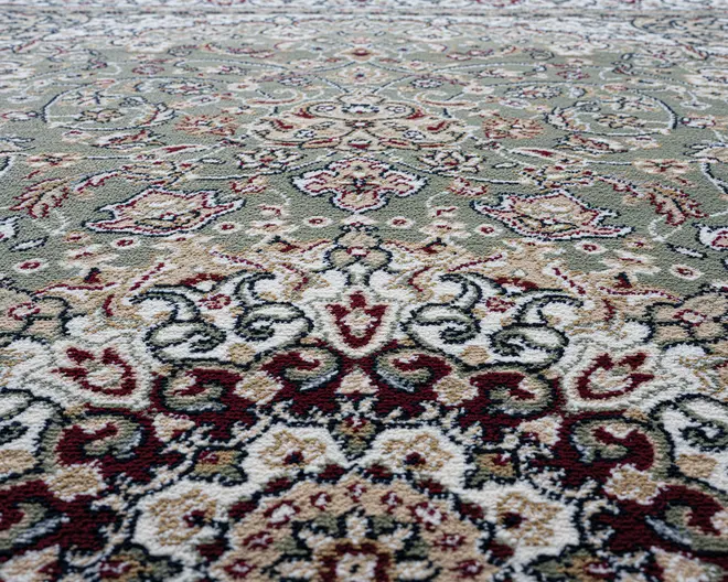 Nærbillede af et traditionelt mønstret tæppe med indviklede blomster- og geometriske motiver. Designet indeholder nuancer af grøn, creme, bordeaux og beige med detaljerede konturer i mørkegrå. Teksturen ser blød og fyldig ud, og mønsteret er symmetrisk, hvilket giver tæppet et klassisk og elegant udtryk.