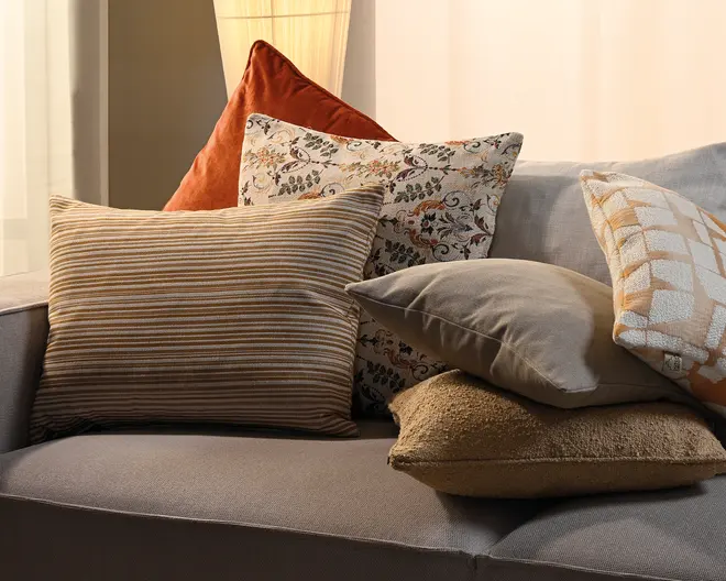 En lagdelt opstilling af pyntepuder på en grå sofa, כולל en stribet pude i beige og brun, en brændt orange velourpude, en cremefarvet pude med blomstermønster, en ensfarvet taupe pude, en tekstureret beige pude og en cremefarvet pude med orange geometrisk mønster.