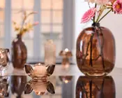 Et nærbillede af en dekorativ opstilling med en bruntonet glasvase med lyserøde og hvide kunstige blomster samt en matchende brun glas fyrfadsstage formet som en blomst. Glasset har en glat, blank overflade og reflekterer lyset på bordpladen. I baggrunden ses flere brune glasvaser og blødt, varmt lys, hvilket skaber en hyggelig og elegant stemning.