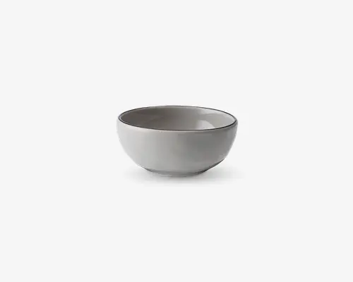 En rund, mellemstor skål i keramik med en glat, blank lysegrå overflade. Skålen har en let udadbuet kant med en tynd, mørk accentlinje langs kanten, hvilket giver et moderne og minimalistisk udtryk. Både inderside og yderside har samme farve, og bunden er flad for stabilitet.