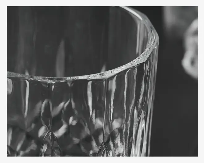 Nærbillede af et klart glas med et facetslebet, geometrisk mønster på den nederste halvdel, hvilket giver en tekstureret og reflekterende overflade. Glassets kant er glat og en smule tyk, og glasset fremstår solidt og elegant.