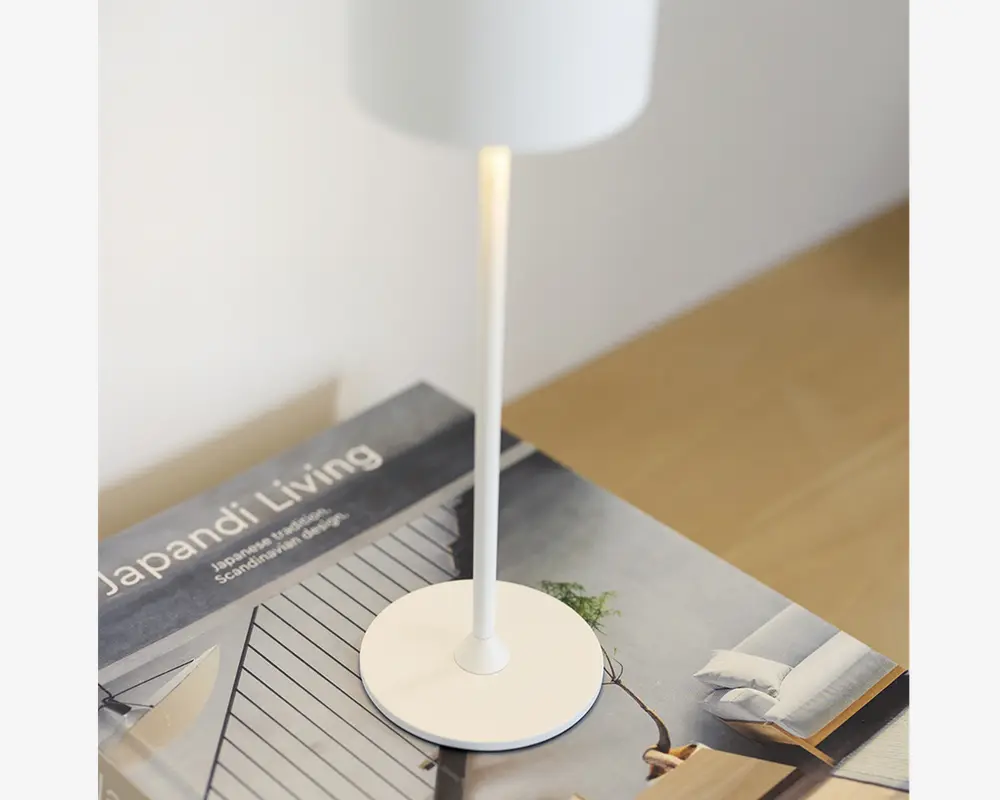 En minimalistisk hvid bordlampe med en slank cylindrisk stang og en rund fod, placeret oven på en bog med titlen 'Japandi Living.' Lampen har en glat mat finish og udsender et blødt, varmt lys nedad. Designet er stilrent og moderne, velegnet til nutidige indretninger.