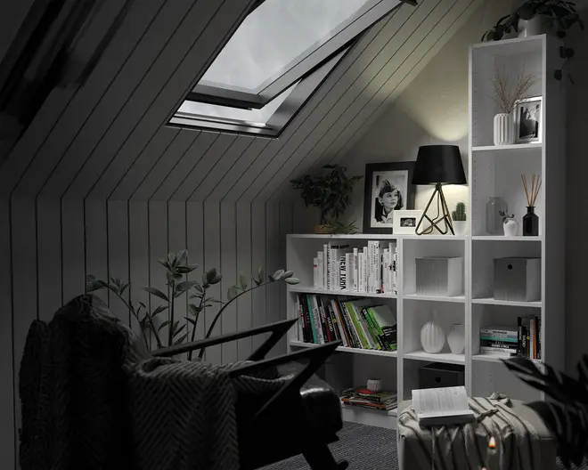 Et moderne, hvidt reolsystem med flere åbne rum, hvor nogle indeholder bøger arrangeret både lodret og vandret, mens andre viser dekorative genstande som vaser, indrammede fotos og små planter. Hylderne er stylet med en blanding af monokrome og naturlige toner, og en sort bordlampe med geometrisk fod står på en af de midterste hylder og kaster et varmt lys. Reolen er placeret op ad en skrå væg under et ovenlysvindue, hvilket bidrager til en hyggelig og moderne stemning.