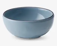 En rund, mellemstor skål af keramik med en glat, blank lyseblå overflade. Skålen har en let tyk kant med en diskret mørk kant og en blødt buet form, der smalner ind mod en lille, stabil fod. Både indersiden og ydersiden har samme farve, hvilket giver et moderne og minimalistisk udtryk.