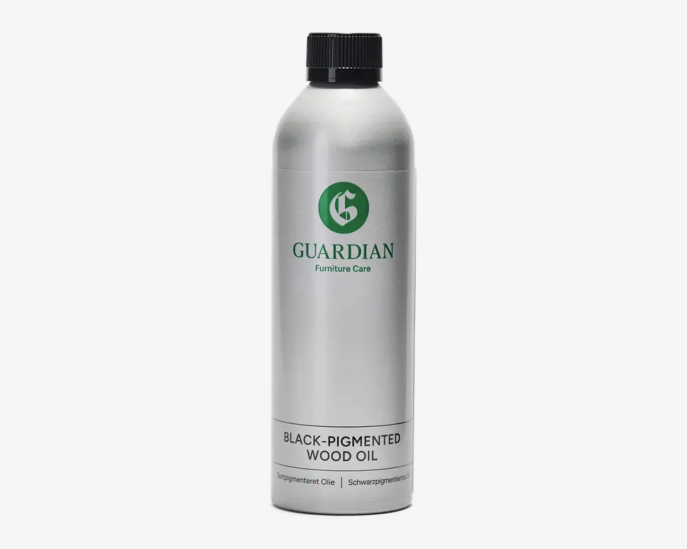 En høj, cylindrisk sølvfarvet metalflaske med sort skruelåg. Etiket med grønt logo og mærket 'Guardian Furniture Care'. Nederst på etiketten står der tydeligt 'Sortpigmenteret Olie'. Flasken har et enkelt og moderne design med tekst på både engelsk og andre sprog, hvilket indikerer, at den er egnet til behandling og beskyttelse af træoverflader med sort pigmentering.