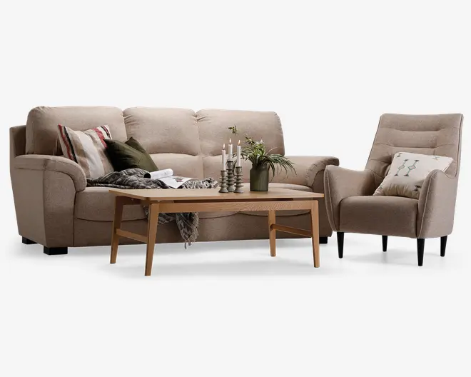 Billedet viser et stuearrangement med en beige polstret sofa og en matchende lænestol med knapdetaljer på ryglænet. Sofaen er pyntet med flere pyntepuder i forskellige farver og mønstre, og et hyggeligt tæppe er lagt over den ene side. Foran siddepladserne står et sofabord i lyst træ, hvorpå der står en grøn vase med friske blomster, tre metalliske lysestager med hvide stearinlys og en lille stak bøger. Lænestolen har også en dekorativ pude med et diskret mønster. Møblerne har sorte ben, og stilen er moderne og indbydende.