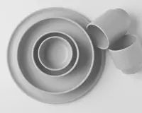 Et sæt minimalistisk service med mat finish i en lys grå farve. Sættet indeholder to tallerkener i forskellige størrelser, en skål og tre kopper. Designet er enkelt og moderne med glatte, afrundede kanter og uden synlige mønstre eller dekorationer.