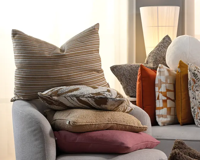 En samling dekorative pyntepuder i forskellige størrelser og teksturer med brede beige- og brunstribede betræk, ensfarvede rust- og sennepsfarvede puder, et creme- og orange geometrisk mønster, en grå tekstureret pude samt andre mønstrede detaljer placeret på polstrede møbler.
