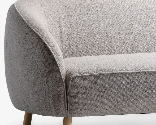 Nærbillede af en moderne lænestol med lysegråt, tekstureret stofbetræk. Armlænet og ryglænet er blødt buede, hvilket giver et indbydende udtryk. Sædehynden er tyk og blød for ekstra komfort. Stolen står på lyse træben, som tilfører et naturligt præg til det moderne design.