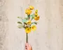 En hånd holder en enkelt kunstig blomsterstilk med flere gule blomsterhoveder og grønne blade. Blomsterne har et lagdelt, realistisk udseende med både knopper og fuldt udsprungne blomster. Stilken er lang og slank, og bladene har en let mat, tekstureret overflade.