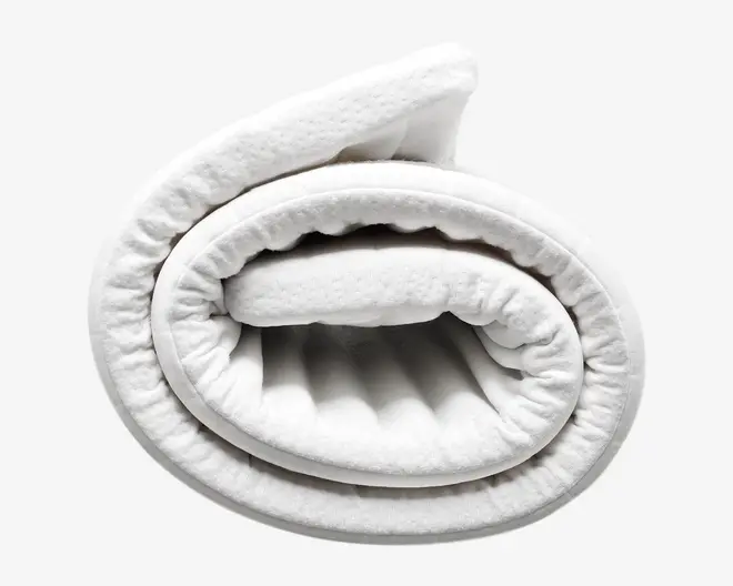 Et nærbillede af en sammenrullet, tyk, hvid topmadras med quiltet overflade. Materialet ser blødt og fyldigt ud med synlig polstring for ekstra komfort. Kanterne er pænt afsluttede, og topmadrassen virker fleksibel og nem at håndtere.