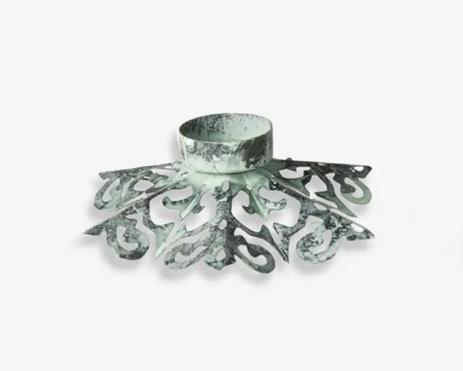 Dekorativ metalholder med et slidt, antikt look. Har detaljerede, symmetriske udskæringer, der stråler ud fra midten og giver et vintage og udsmykket udtryk. Den centrale, cirkulære del er designet til at holde en genstand sikkert, mens den brede base giver stabilitet.