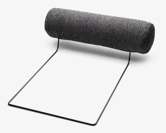 En cylindrisk, mørkegrå pude er fastgjort til en minimalistisk sort metalramme. Puden er betrukket med et blødt, tekstureret stof. Rammen er åben og vinklet, designet til at holde puden i en hævet position. Det samlede design er moderne og funktionelt, sandsynligvis beregnet til ergonomisk støtte, såsom en nakkestøtte eller hovedpude til en stol eller sofa.