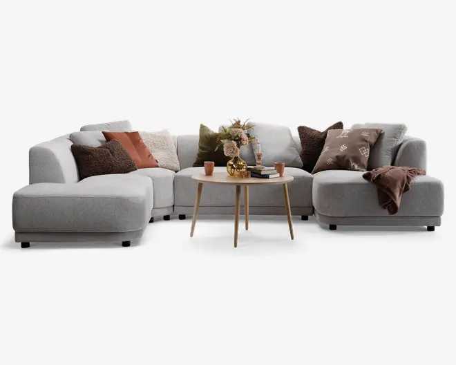 En moderne, L-formet hjørnesofa betrukket med lysegråt stof. Sofaen har brede, afrundede armlæn og bløde sædehynder. Den er pyntet med forskellige pyntepuder i jordfarver, herunder brun, olivengrøn, rust og creme, nogle med tekstur eller mønster. Et blødt brunt tæppe er lagt over det ene armlæn. Foran sofaen står et rundt sofabord i lyst træ med en vase med blomster, en dekorativ gylden genstand, flere bøger og et par små kopper.