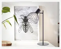 En moderne bordlampe med en slank, mat sort metalstand og rund fod. Lampen har en unik lampeskærm af metaltråd, som gør pæren synlig og giver et industrielt udtryk. Lampens hoved kan justeres, så lyset kan rettes. Lampen står på en hvid overflade ved siden af et indrammet kunstværk og en grøn plante.