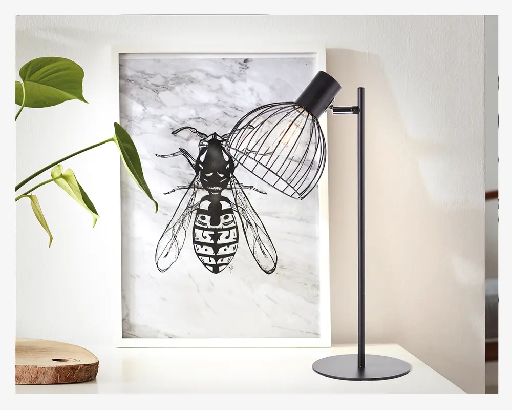 En moderne bordlampe med en slank, mat sort metalstand og rund fod. Lampen har en unik lampeskærm af metaltråd, som gør pæren synlig og giver et industrielt udtryk. Lampens hoved kan justeres, så lyset kan rettes. Lampen står på en hvid overflade ved siden af et indrammet kunstværk og en grøn plante.