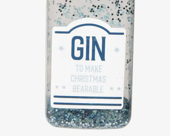Et nærbillede af en gennemsigtig flaske dekoreret med sølv- og blå glimmer. Flasken har en hvid etiket med blå tekst, hvor der står 'GIN' og mindre tekst nedenunder, der siger 'TO MAKE CHRISTMAS BEARABLE.' Etiketten er pyntet med små stjerner og blå linjer for et festligt udtryk.