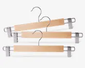 Sæt med tre bøjler i træ med naturfinish, hver med en solid metalkrog og to justerbare metalclips med plastikgreb, der sikrer, at bukser, nederdele eller andre beklædningsgenstande holdes fast uden at glide eller efterlade mærker.