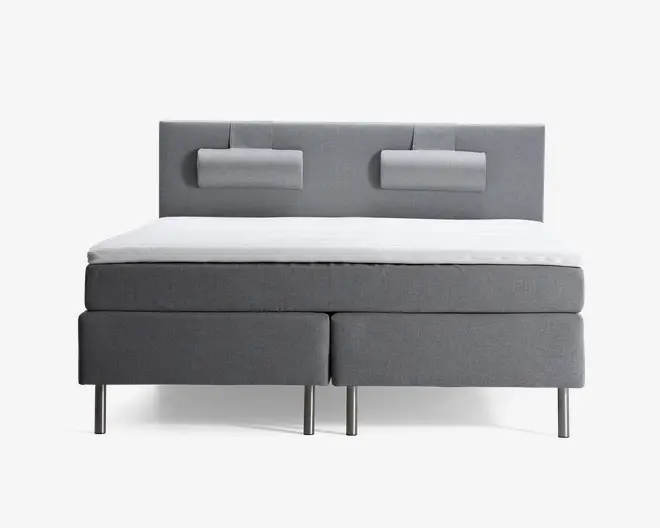 En moderne dobbeltseng med gråt polstret stel og sengegavl. Sengegavlen har to rektangulære, polstrede puder fastgjort for ekstra komfort. Sengen står på seks slanke, metalben, og madrassen er dækket af en hvid topmadras eller madrasbeskytter. Designet er minimalistisk og moderne.