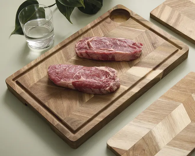 Rektangulært skærebræt i træ med et geometrisk chevron-mønster, en saftrille langs kanten og et rundt udskåret hul i det ene hjørne. Brættet har en glat, poleret overflade og naturlige variationer i træets årer.