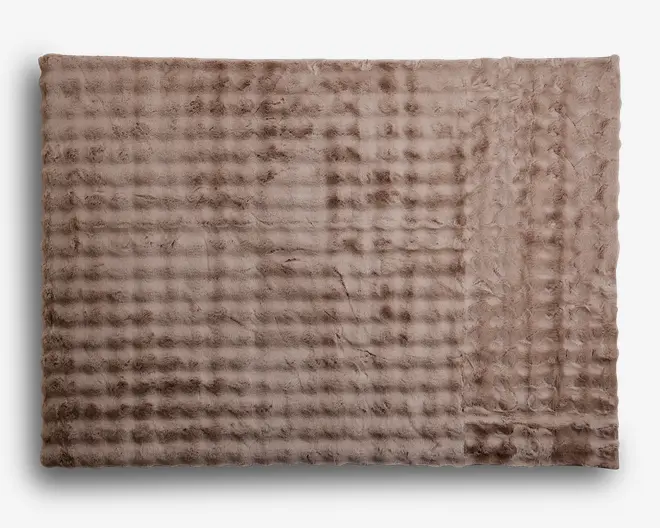 Blød, rektangulær plaid med en plysset, kunstpels-lignende tekstur i en lys taupe farve. Overfladen har et diskret, ternet mønster, som giver et hyggeligt og indbydende udtryk. Kanterne er pænt afsluttede, og materialet fremstår tykt og varmt, perfekt til komfort og varme.