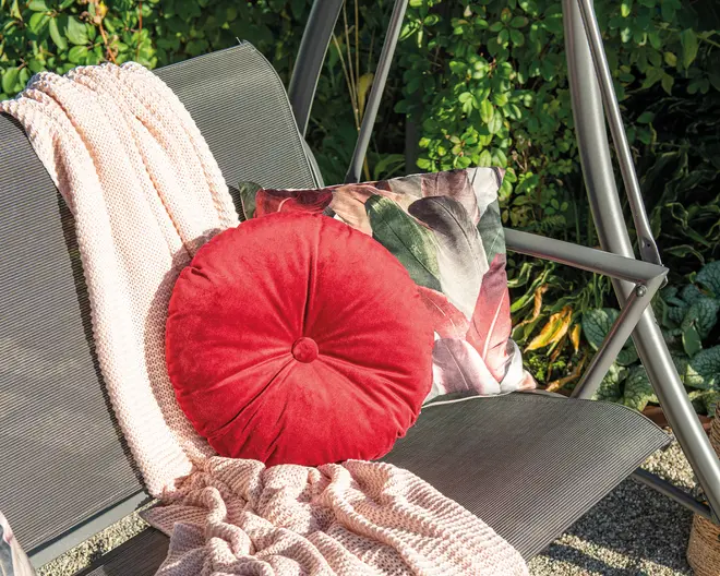 En rund, blød rød pude med en knapdetalje i midten ligger på en bænk. Bagved er der en firkantet pude med botanisk print i grønne og lyserøde nuancer. Et blødt, groftstrikket lyserødt tæppe er lagt over bænken og giver et hyggeligt udtryk. Bænken er lavet af metalslats og står udendørs omgivet af frodigt grønt løv.