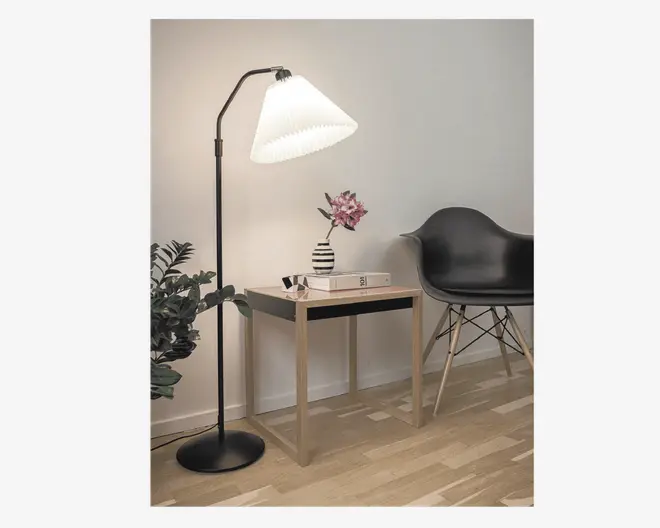 Et moderne, minimalistisk sidebord med en lys træfinish og enkle firkantede ben. Bordet er dekoreret med en sort og hvidstribet vase med en lyserød kunstig blomst samt to stablede bøger. Opsætningen inkluderer også en sort gulvlampe med en hvid plisseret lampeskærm, en sort stol i formstøbt plast med træben og en grøn, bladfyldt plante i baggrunden. Rummet har lyst trægulv og en ensfarvet hvid væg, hvilket skaber et rent og moderne udtryk.
