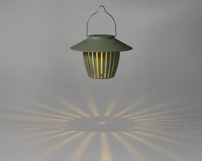En hængende lanterne med mat grøn finish og en bred, skrånende top. Lanternen har lodrette lameller rundt om kroppen, som lader varmt lys skinne igennem og skaber et dekorativt stjernemønster på overfladen nedenunder. Den har et metalhåndtag til nem transport eller ophængning.