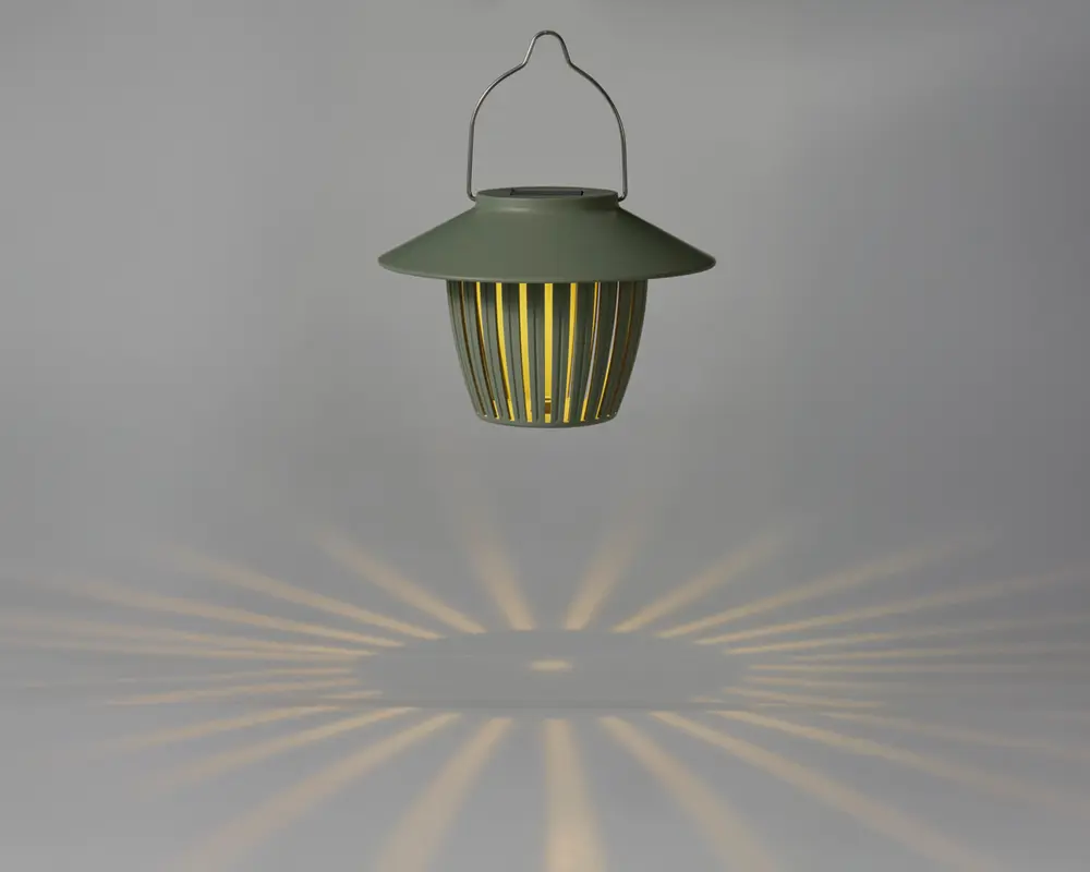 En hængende lanterne med mat grøn finish og en bred, skrånende top. Lanternen har lodrette lameller rundt om kroppen, som lader varmt lys skinne igennem og skaber et dekorativt stjernemønster på overfladen nedenunder. Den har et metalhåndtag til nem transport eller ophængning.