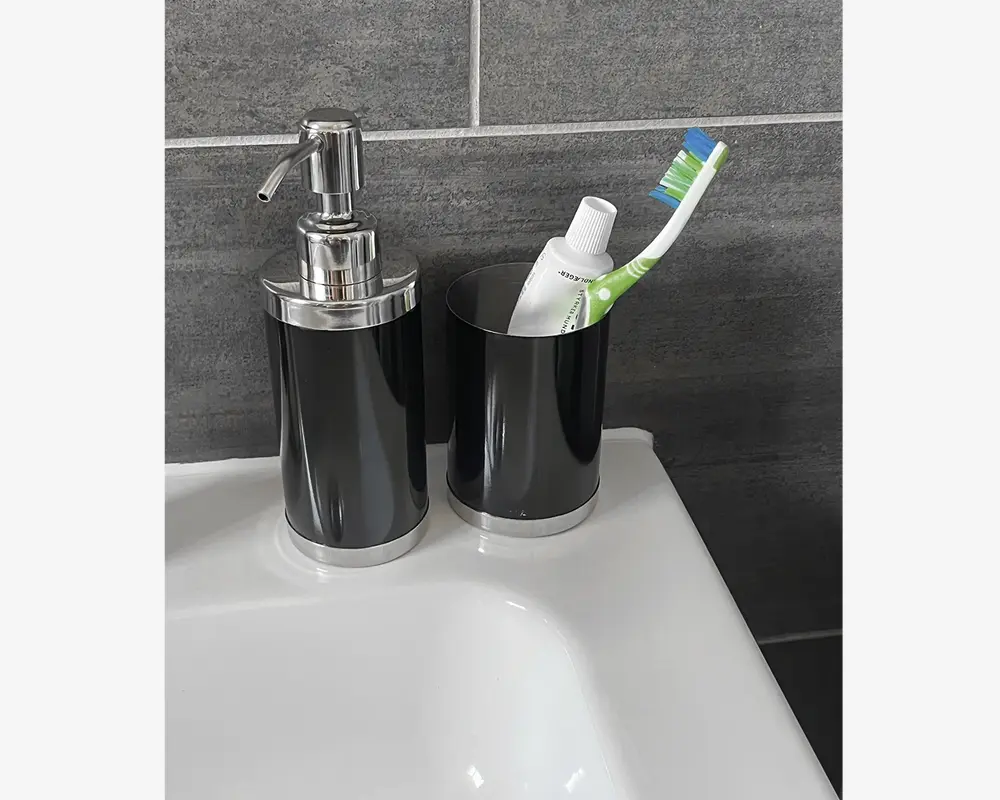 Et moderne badeværelsessæt på bordet bestående af en sort og sølv pumpe-dispenser med metaltop, velegnet til flydende sæbe eller lotion, samt en matchende sort og sølv tandkrus, der indeholder en tandbørste og tandpasta. Begge dele har en glansfuld overflade og et minimalistisk design, som passer til de mørke fliser i baggrunden.