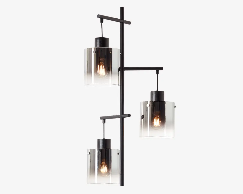 Et moderne vægmonteret armatur med tre cylindriske glasskærme, der har en farveovergang fra klart glas i bunden til røgfarvet sort i toppen. Hver skærm indeholder en synlig glødepære, og skærmene er fastgjort til en slank, sort metalramme med vandrette arme i forskellige højder for et forskudt, geometrisk udtryk.