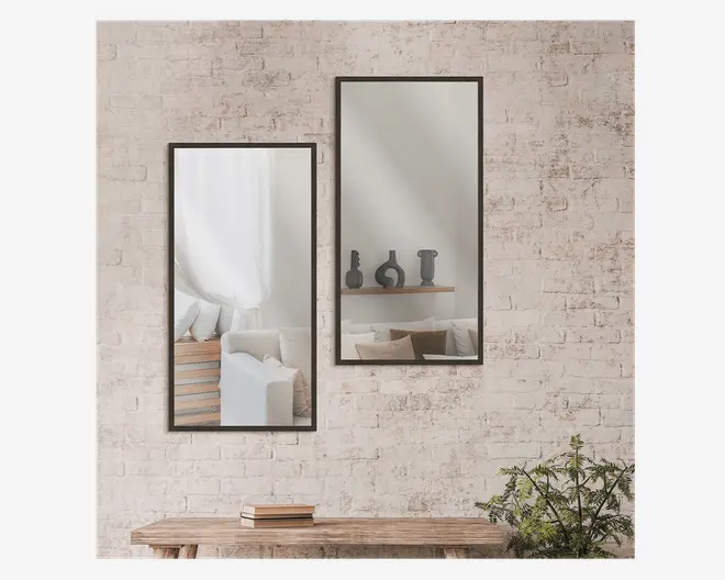 To rektangulære vægspejle med slanke sorte metalrammer er monteret side om side på en lys murstensvæg. Spejlene har et moderne, minimalistisk design og reflekterer et hyggeligt interiør med møbler i neutrale toner, dekorative puder og en træhylde med skulpturelle vaser. Spejlene er placeret vertikalt og giver rummet en følelse af dybde og lys.