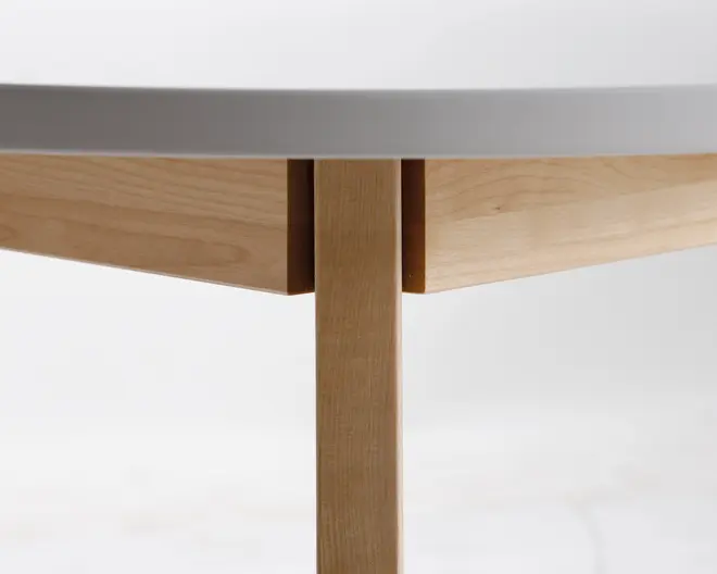 Nærbillede af et bordhjørne, der viser et glat, lyst træben og sarg med rene, lige linjer. Bordpladen har en mat, lysegrå overflade med en afrundet kant. Samlingerne er præcise og fremhæver det gode håndværk og det moderne, minimalistiske design.